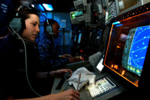cryptologic technician sits at desk with headset on and faces computer monitor with a radar screen in a dimly lit room with another sailor working beside her